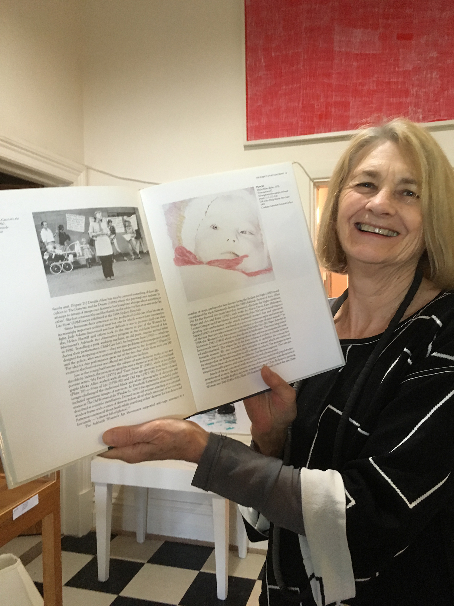 Photograph of a person with short blonde hair, smiling and holding open a book to the camera